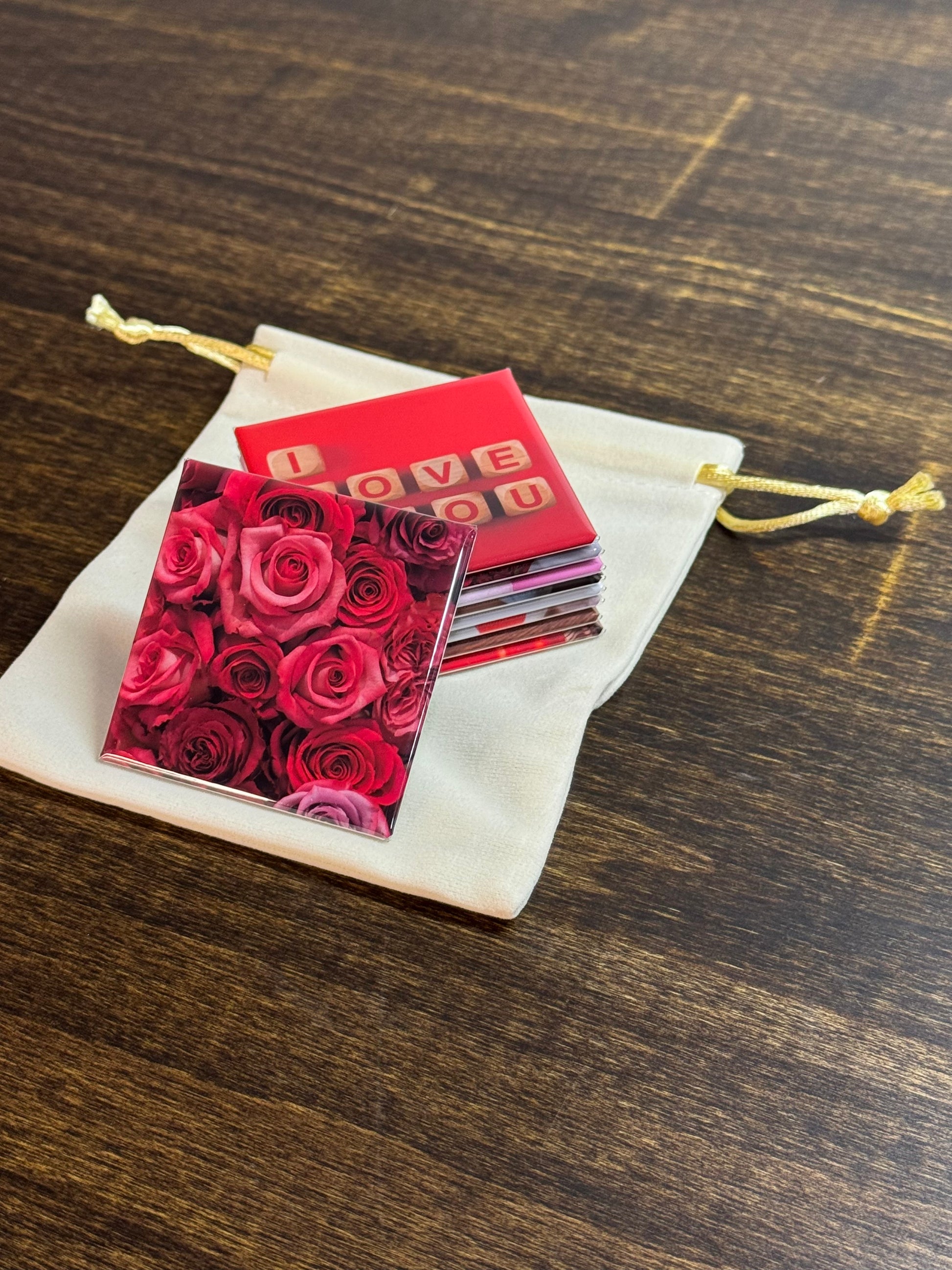 Set of 2x2 inch square magnets with vibrant, red floral design and 'I Love You' text on a wooden surface. The set of magnets is sitting on a cream colored, velvet baggie that is perfect for gifting.