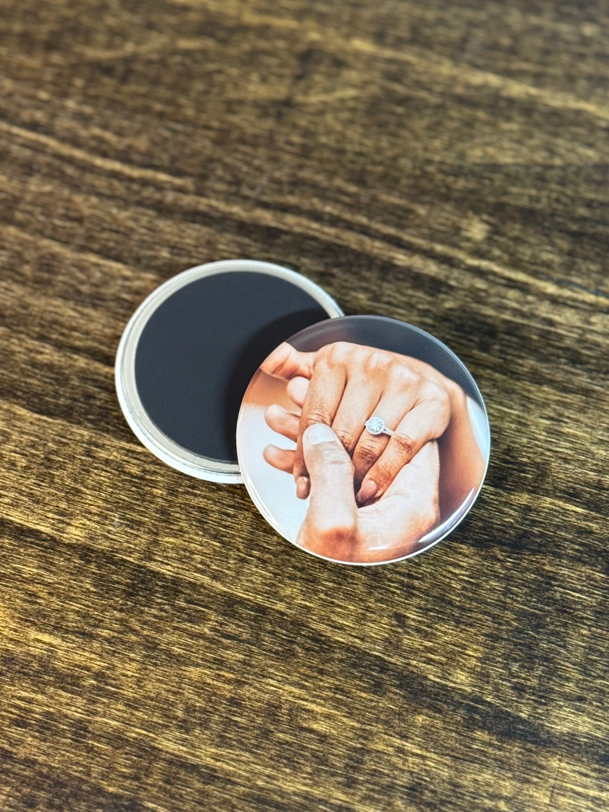 Two round magnets, one flipped to show the back of the magnet. The other magnet shows two hands holding, and one of the hands is showing an engagement ring. 