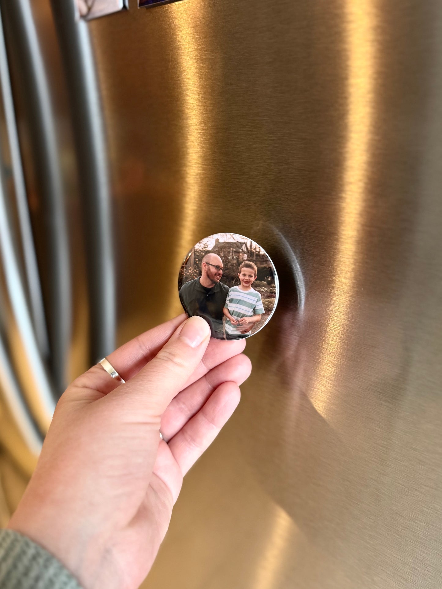 A round photo magnet being held in a hand by a refrigerator. The photo on the magnet shows a father and son. 