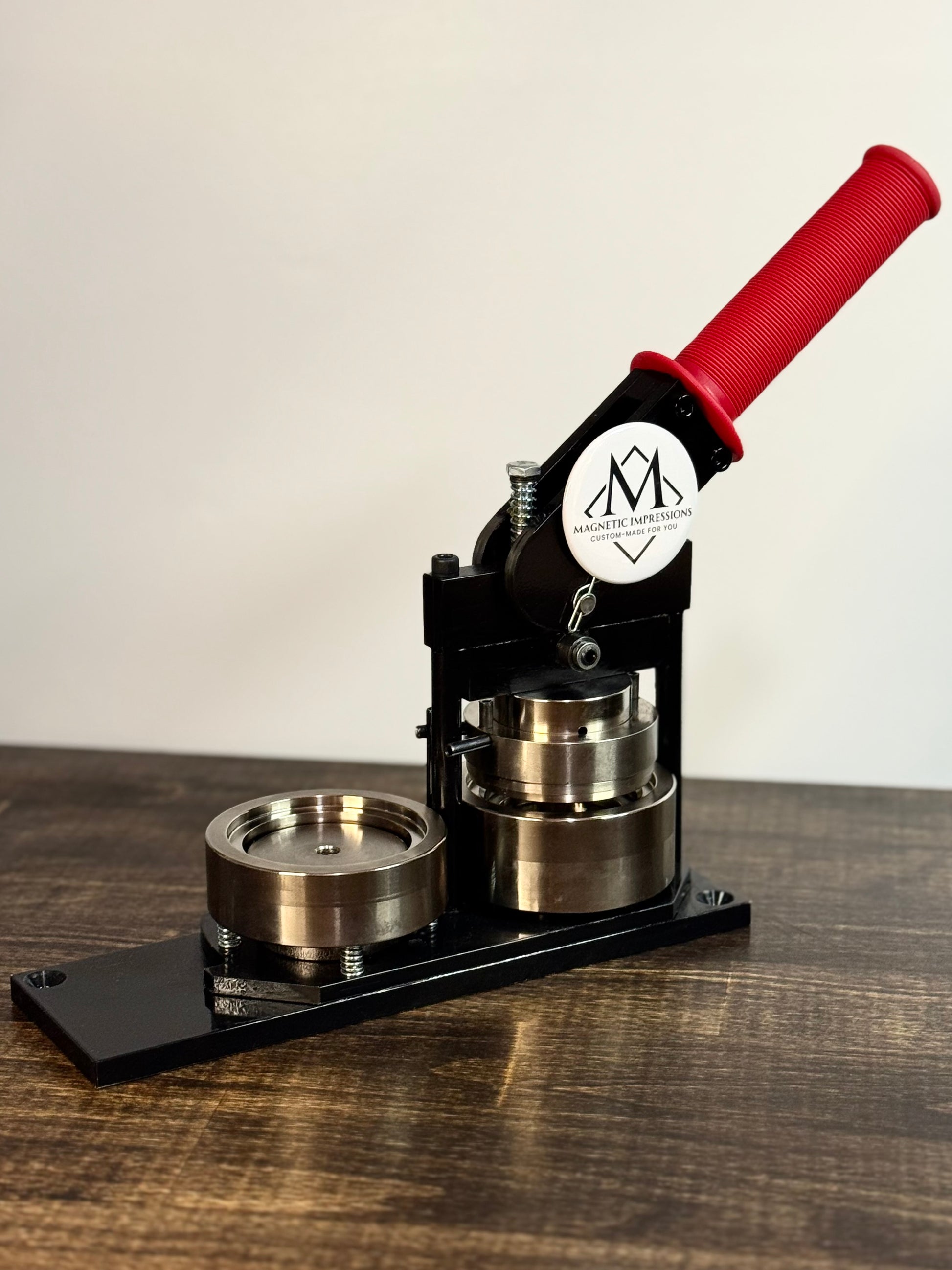 Professional-grade magnet press with red handle on a wooden table. Round magnet on the machine reads "Magnetic Impressions Custom-Made for You"