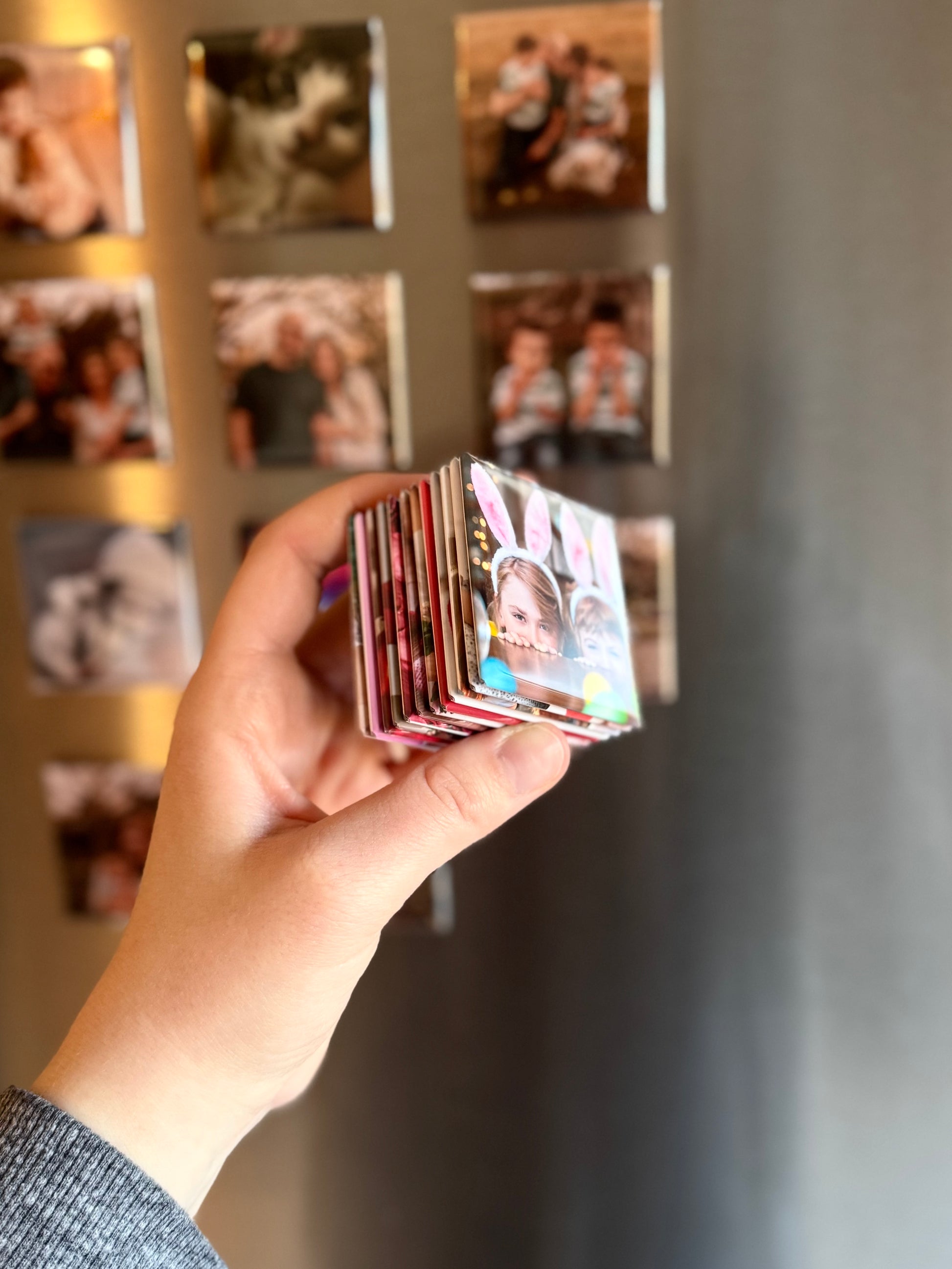 Hand holding a stack of 2x2 inch custom photo magnets, showing the thickness and glossy square finish, with personalized images layered together and photo magnets displayed on a refrigerator in the background.