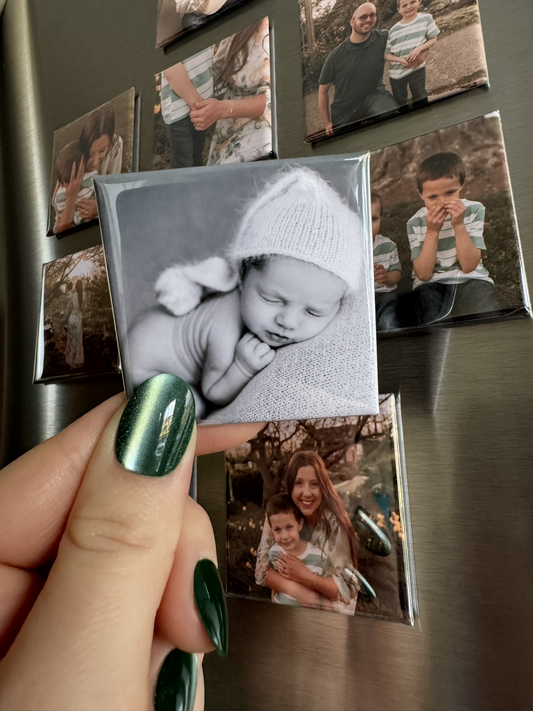 Custom 2x2 inch square photo magnet held in hand on a refrigerator, showing a personalized baby photo and family photos in the background
