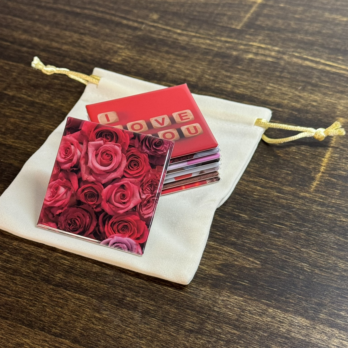 A set of nine square magnets stacked on a cream colored, velvet baggie. One of the square magnets shows vibrant, red roses, and the other magnet stacked on top reads "I love you"
The image shows the glossy shine of the magnets. 