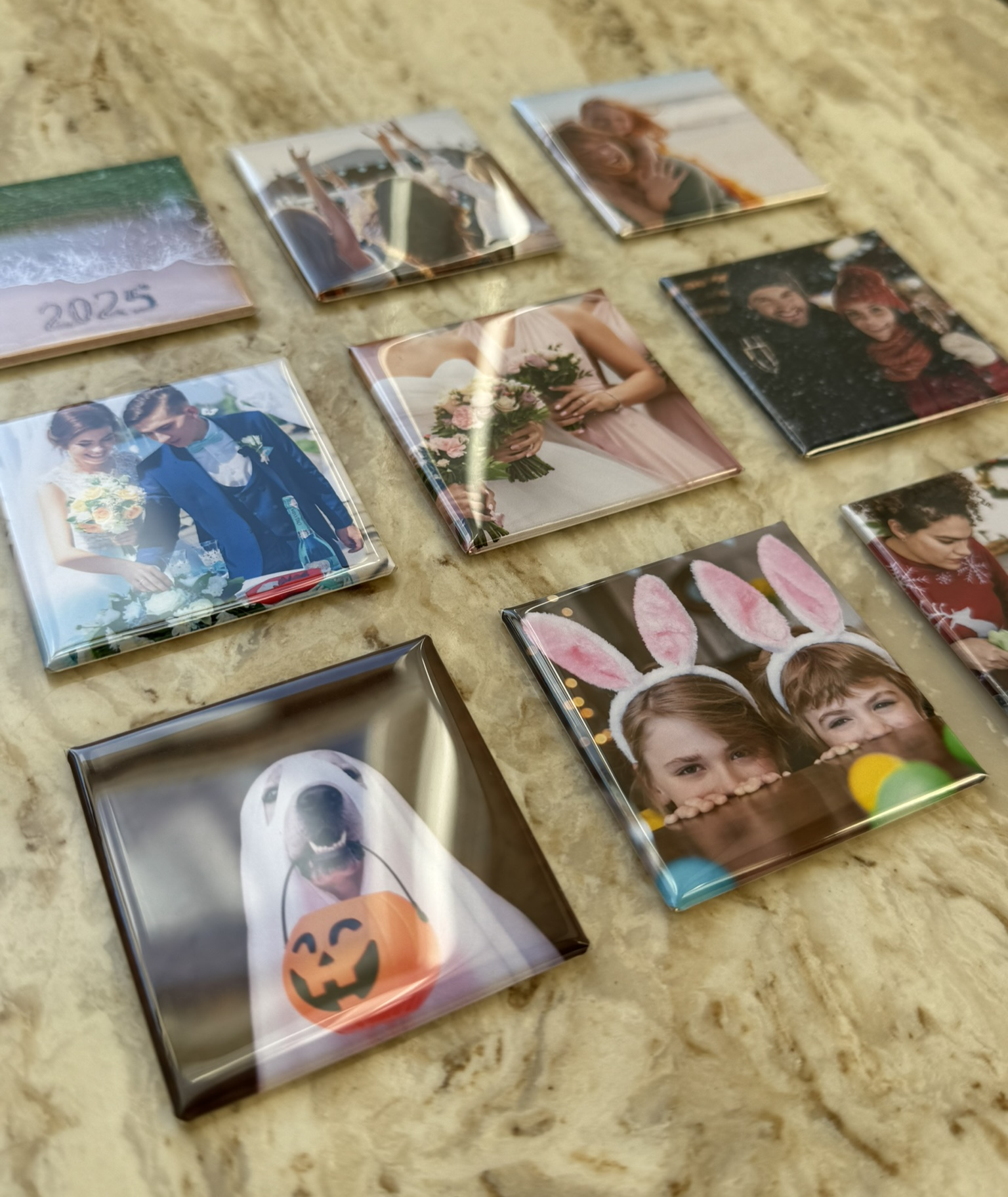 Assorted 2x2 inch custom photo magnets displayed on a countertop, featuring personalized images including weddings, family photos, holidays, and pets, shown in a glossy square finish