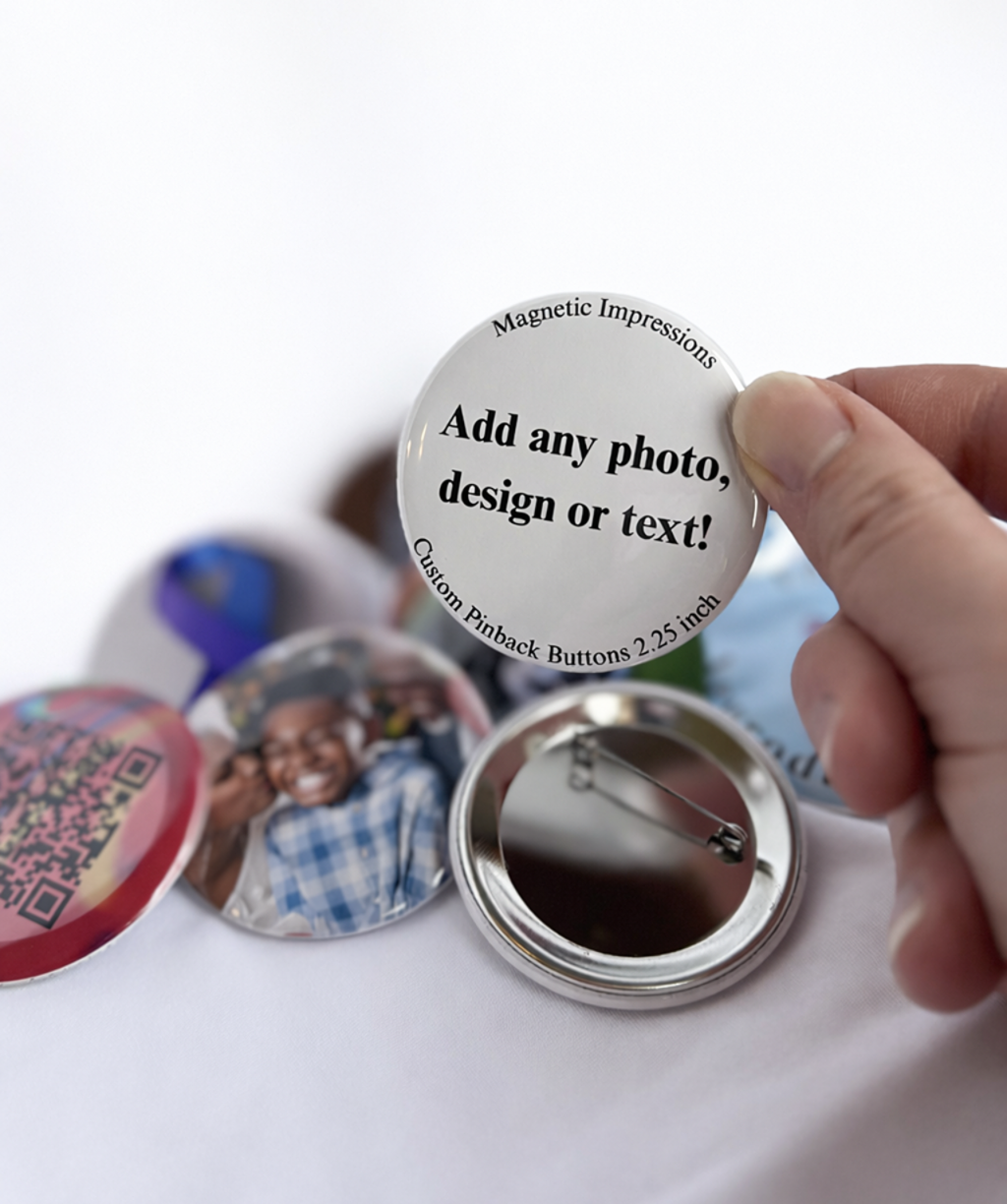 Hand holding a 2.25 inch round customizable button with text 'Add any photo, design or text!' on a blurred background. One of the buttons is turned around to show the metal pinback.
