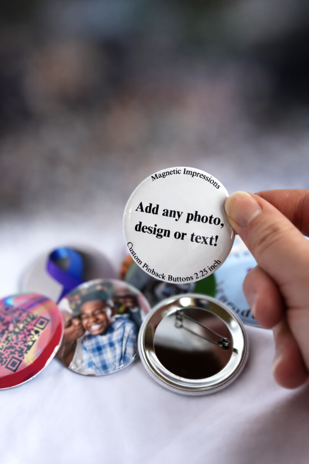 Hand holding a 2.25 inch round customizable button with text 'Add any photo, design or text!' on a blurred background. One of the buttons is turned around to show the metal pinback.